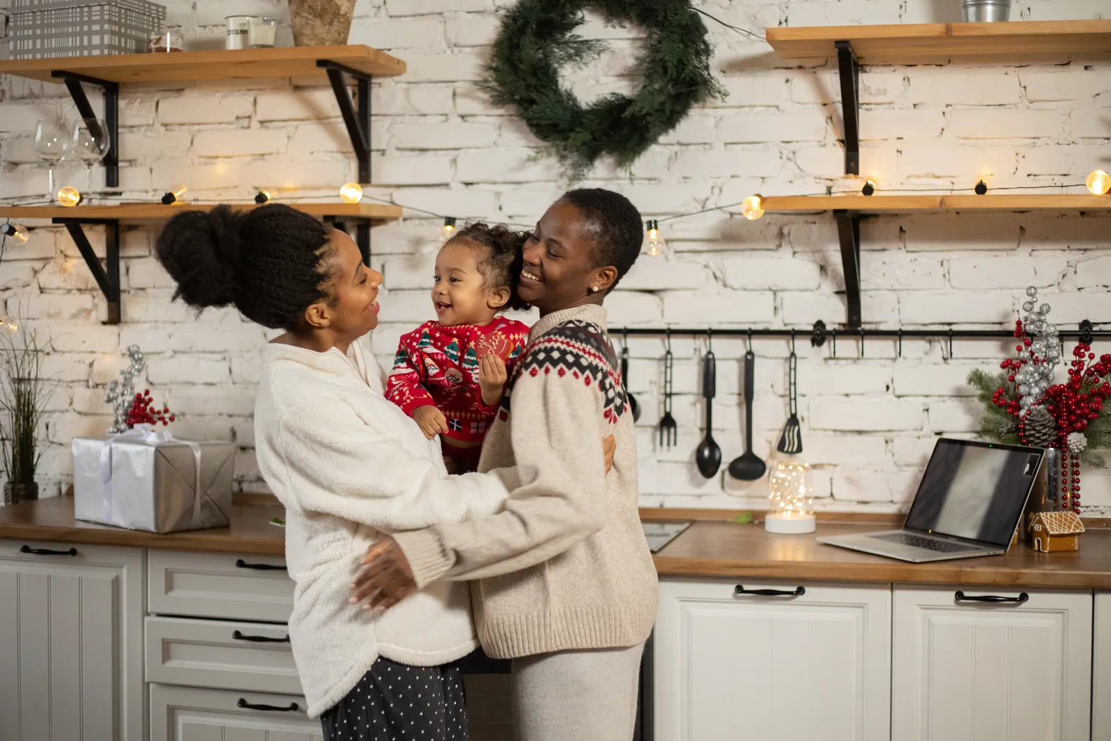 Two women hug, a laughing baby held between them, in a kitchen strung with fairy lights.