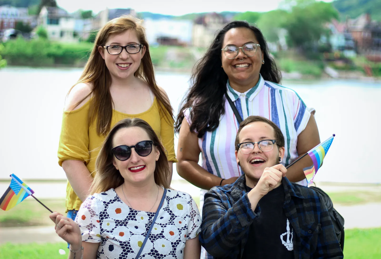 Four friends smile together outdoors by a river, two of them waving small flags.