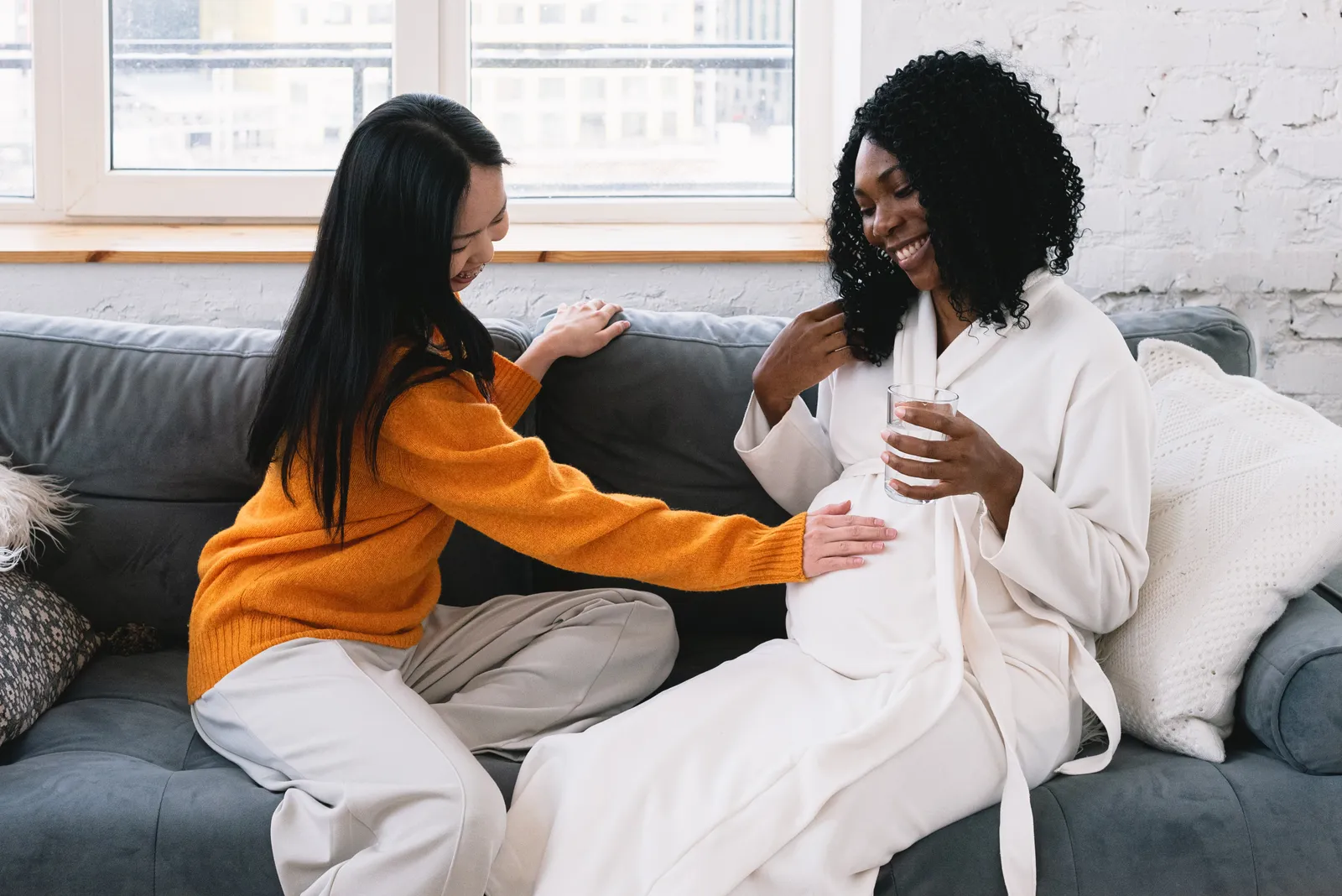 A woman rests her hand on her pregnant friend's belly as they laugh together on a sofa.