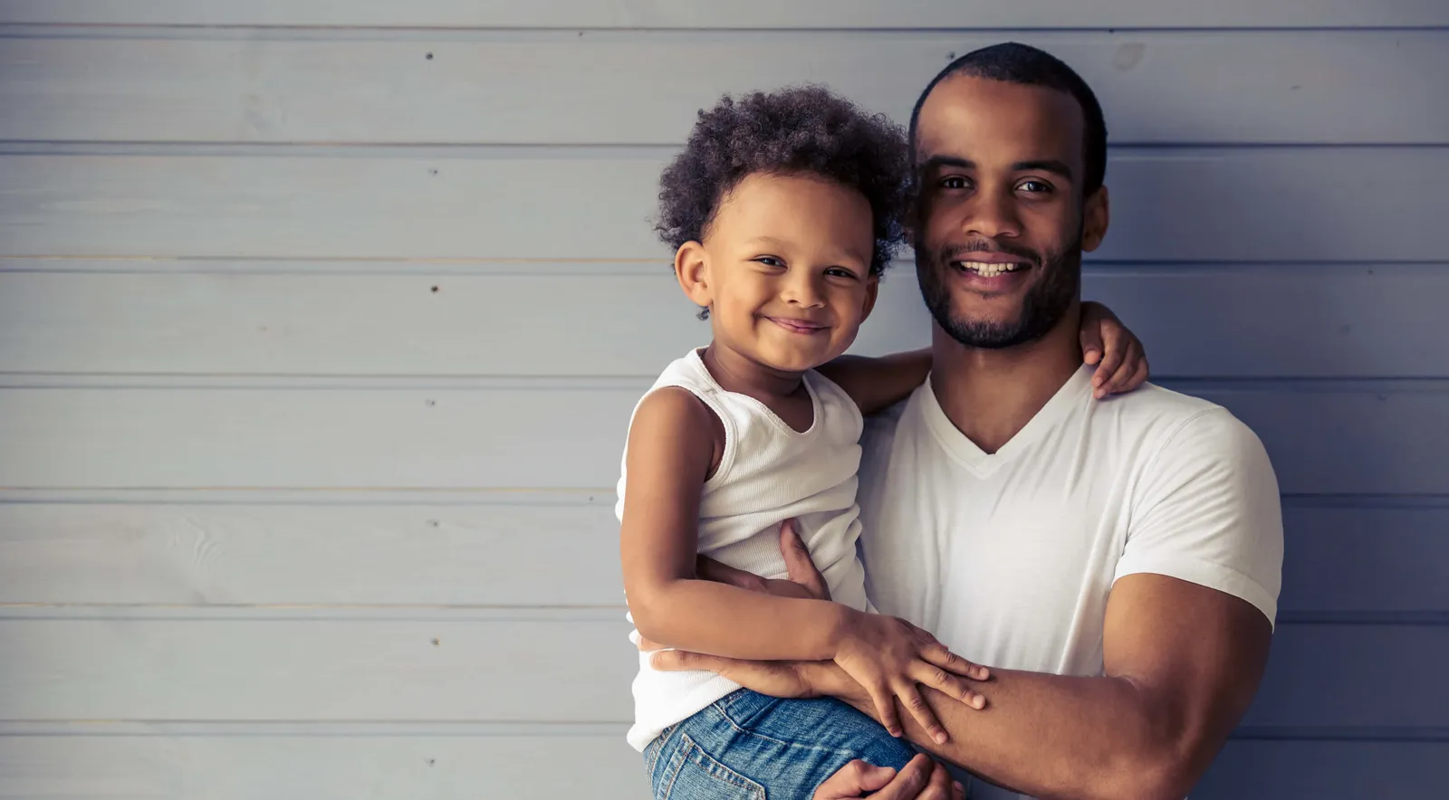 A smiling man holds a little boy on his hip in front of a pale wooden wall.