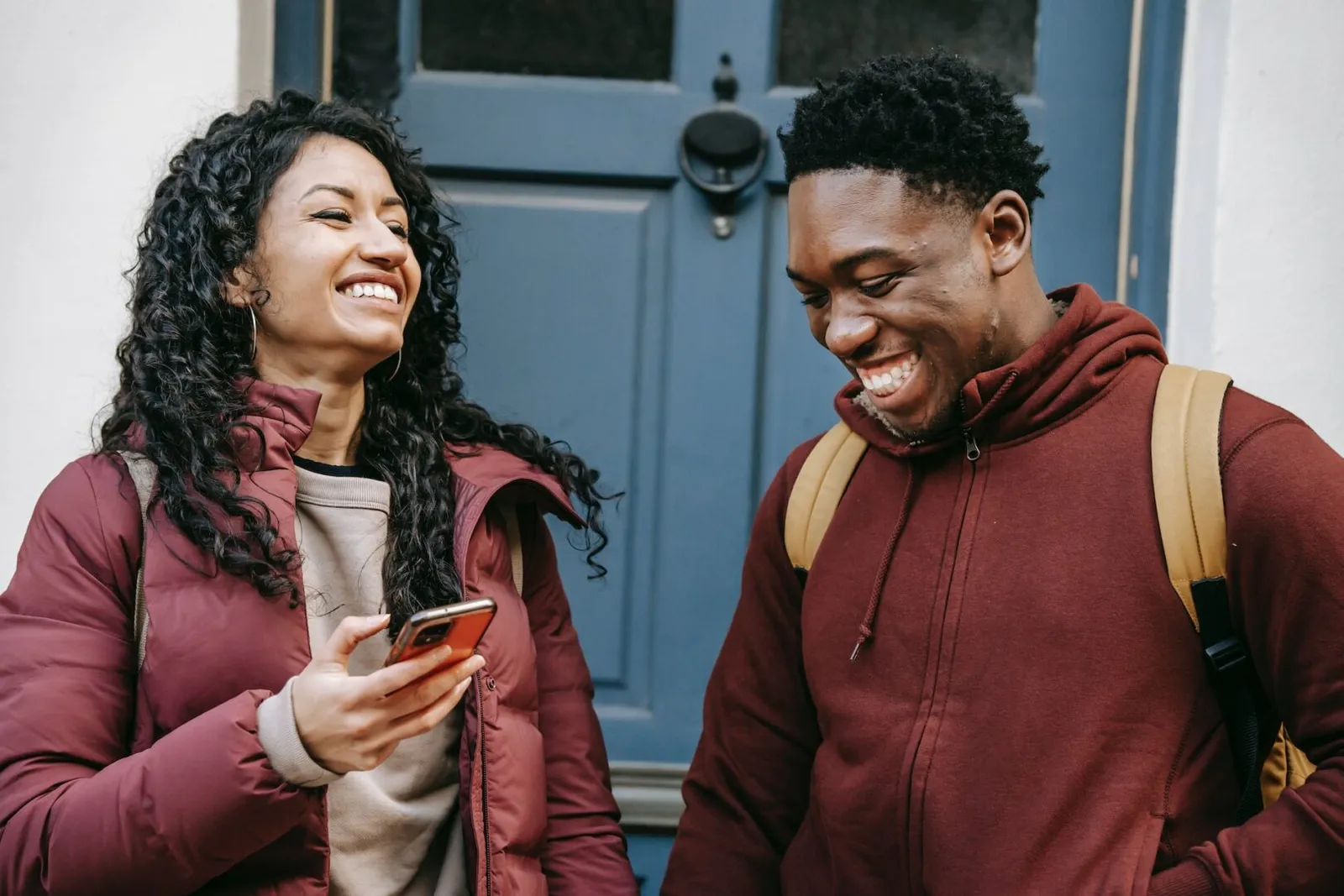 A man and a woman laugh together on a doorstep, her phone in her hand.